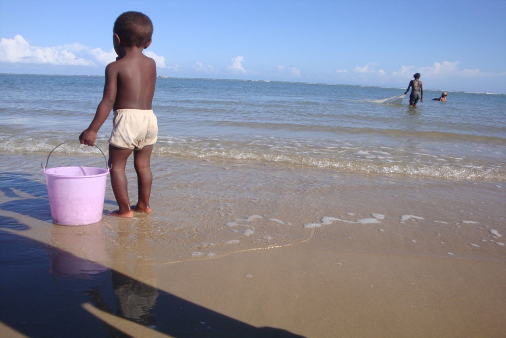 A small boy holds onto a bucket as he wades on the beach in Anosikely, a neighbourhood in Morondava, Madagascar. Behind him, women use nets to fish. Photo: UNICEF/UNI72915/de Paul